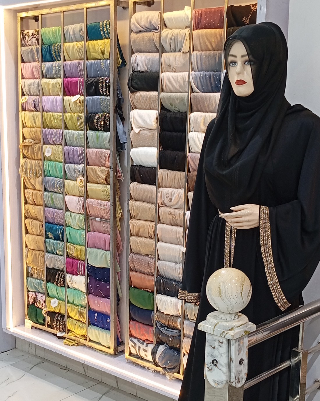 Mannequin wearing a black embroidered abaya with gold thread borderwork beside a rack of hijabs